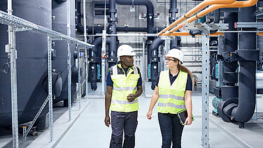 Two Leadec employee wearing safety vests and helmets walk through a technical area.